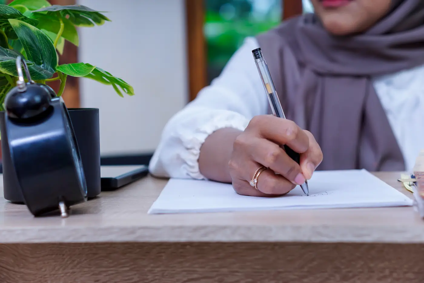 Vrouw met hoofddoek die met een pen op een vel papier schrijft aan een houten bureau